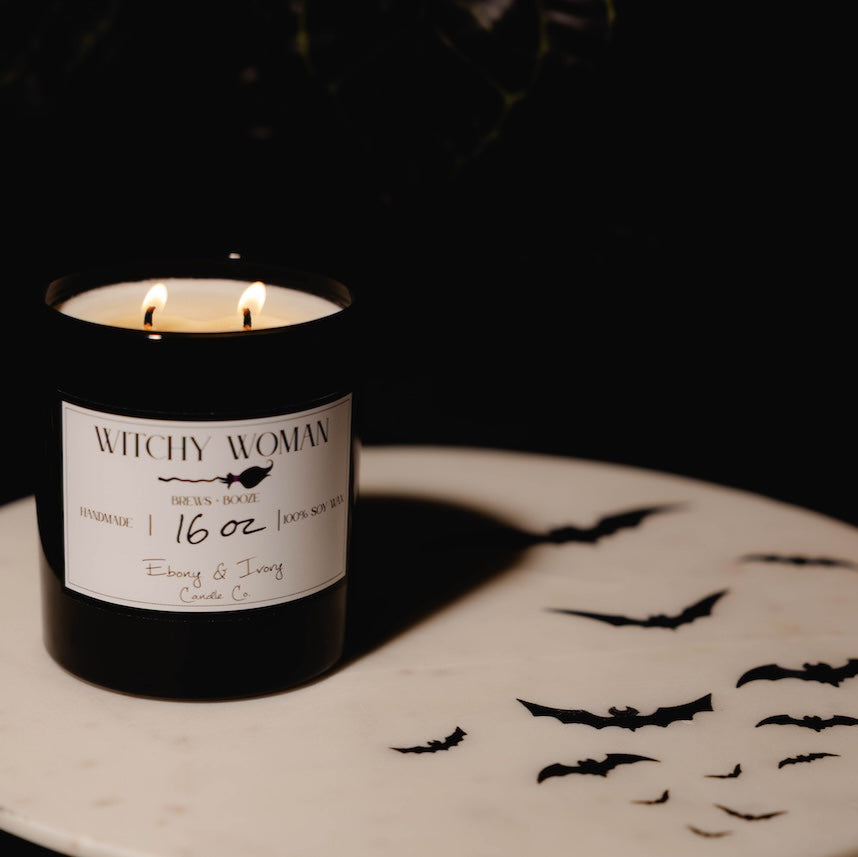 black 16oz soy candle with a white label named "witchy woman" on a white table with black bats
