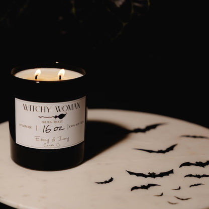 black 16oz soy candle with a white label named "witchy woman" on a white table with black bats