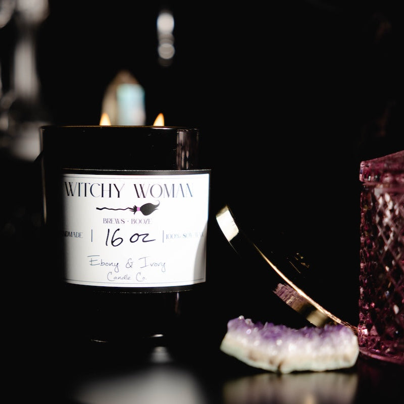 Black 16oz soy candle with a white label named "witchy woman" on a dark background with witchy decor