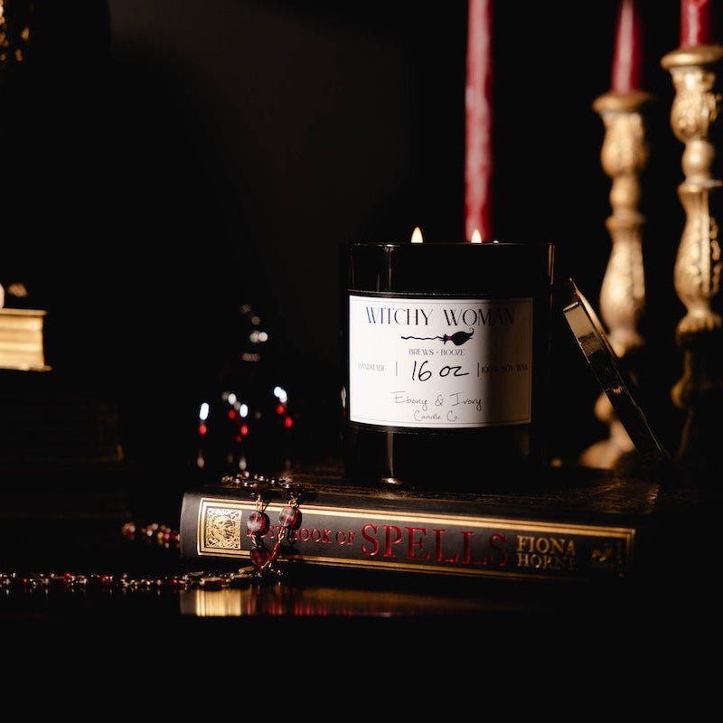 Black 16oz soy candle with a white label named "Witchy Woman" sitting on a spell book with red, black, and gold romantic decor.