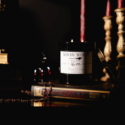Black 16oz soy candle with a white label named "Witchy Woman" sitting on a spell book with red, black, and gold romantic decor.