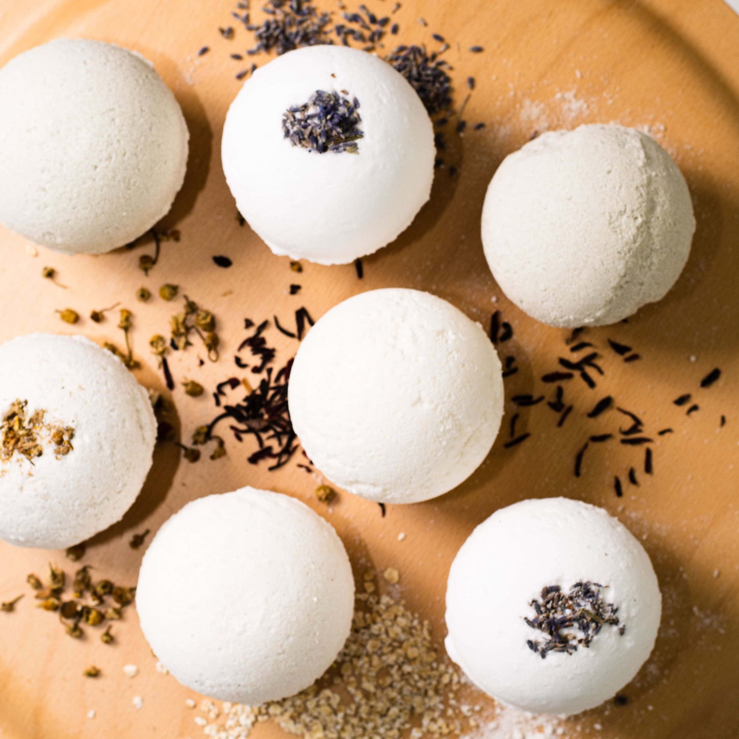 7 white bath bombs, some made with pumpkin powder, some made with lavender buds, some made with green tea powder, and some made with chamomile buds, all of them sitting on a wooden tray, made by Ebony & Ivory Candle Co in Lethbridge, Alberta, Canada