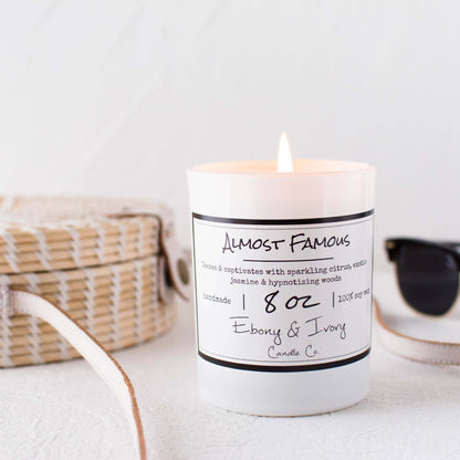 A white 8oz jar soy candle with a white label that reads "Almost Famous" sitting on a clean white table with a beachy wicker bag and sunglasses behind it, made by Ebony & Ivory Candle Co in Lethbridge, Alberta, Canada