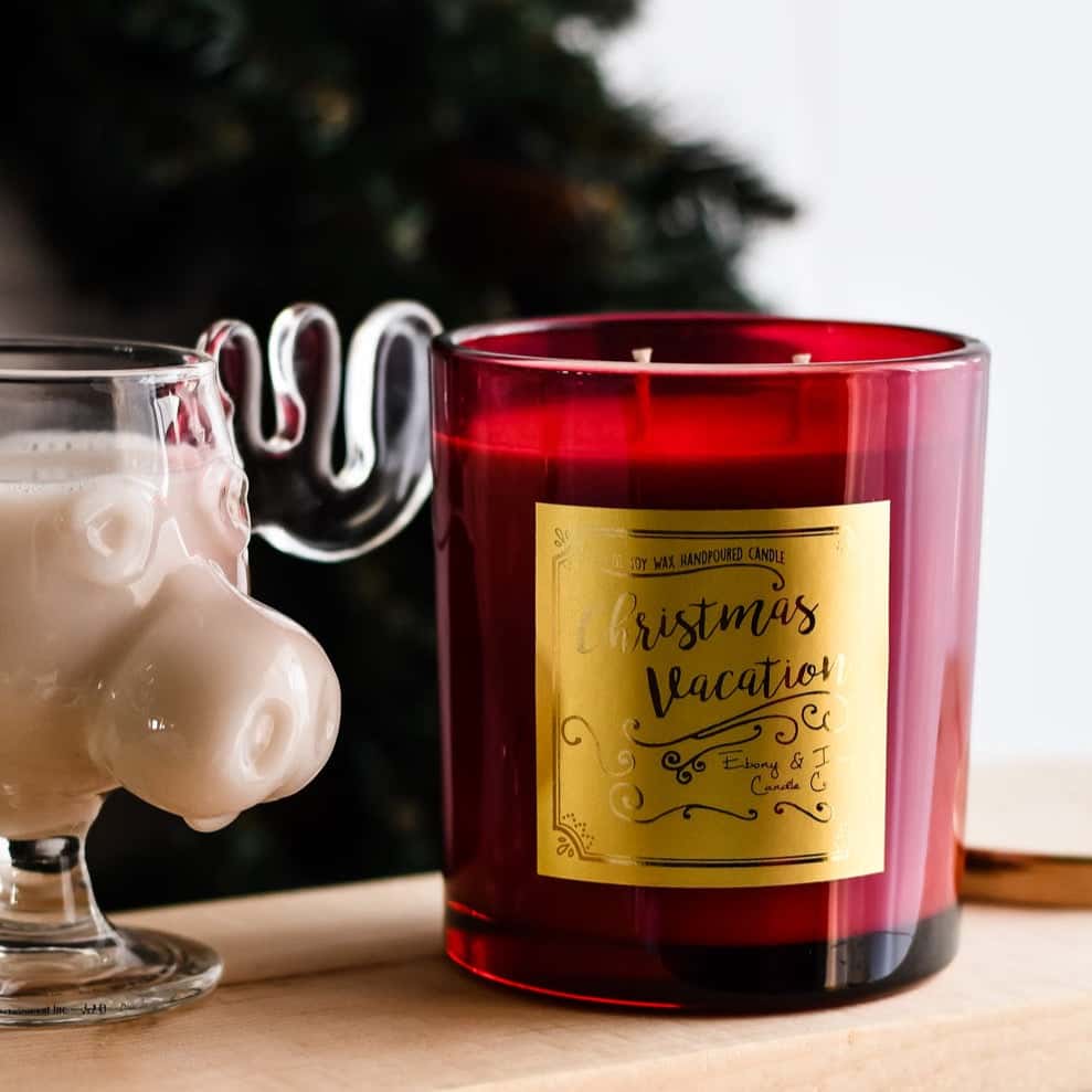 A red 16oz soy christmas candle with a gold label named "christmas vacation" sitting next to a moose glass full of holiday eggnog