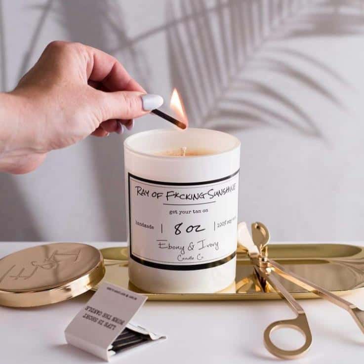 A white 8oz soy candle labelled Ray of F*cking Sunshine, sitting on a gold tray with gold wick trimmers and a gold lid, being lit by a woman. Made in Lethbridge Alberta Canada