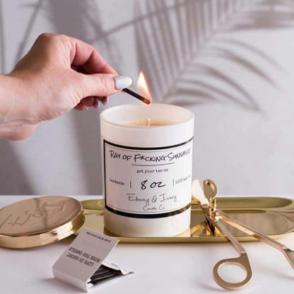 A white 8oz soy candle labelled Ray of F*cking Sunshine, sitting on a gold tray with gold wick trimmers and a gold lid, being lit by a woman. Made in Lethbridge Alberta Canada
