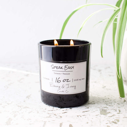 A black jar 16oz soy candle with a gold lid and a white label named Speak Easy, on a clean granite counter top with a potted plant beside it, made by Ebony & Ivory Candle Co in Lethbridge, Alberta, Canada