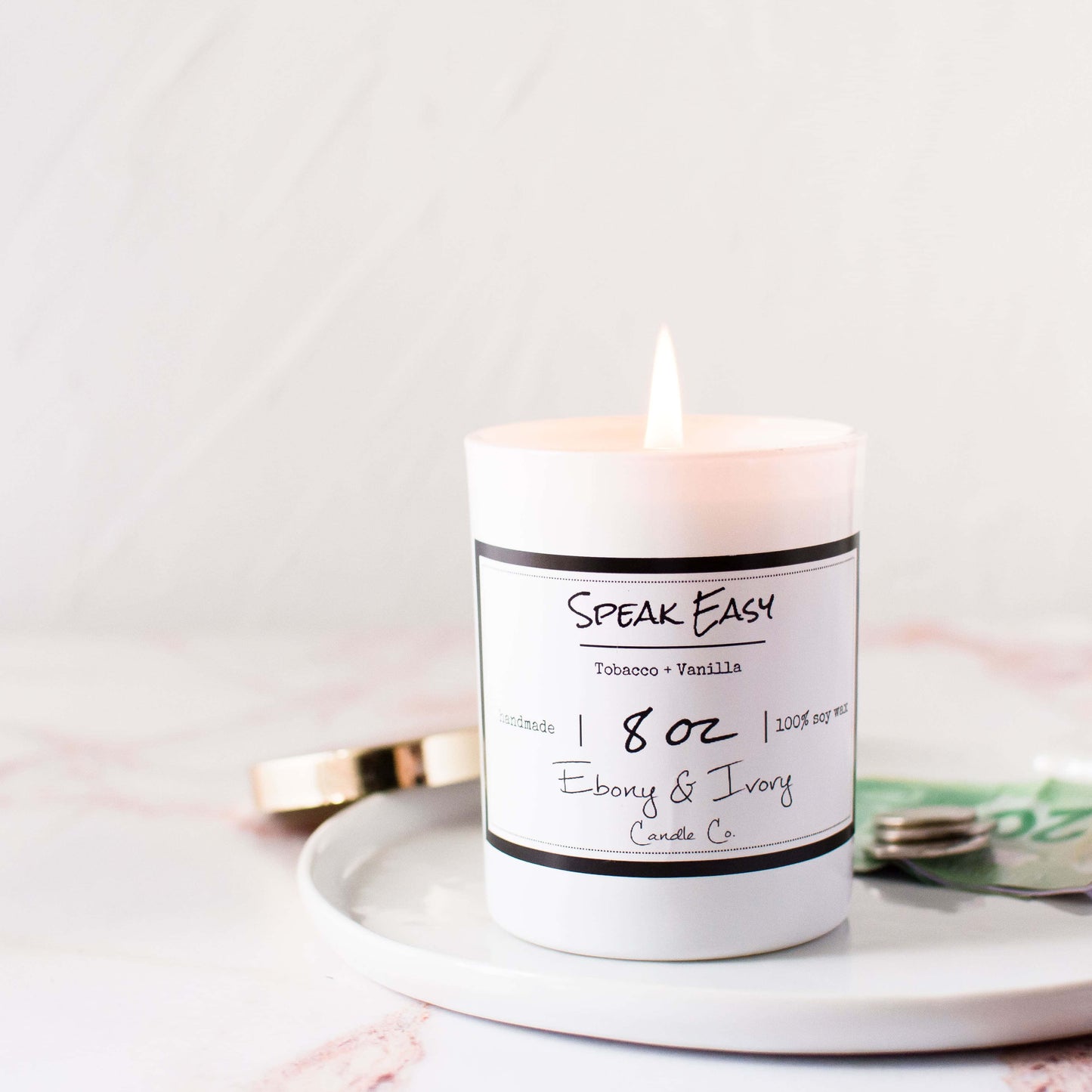 A white jar 8oz soy candle with a gold lid and a white label named Speak Easy, on a white tray on top of a marble counter top, made by Ebony & Ivory Candle Co in Lethbridge, Alberta, Canada