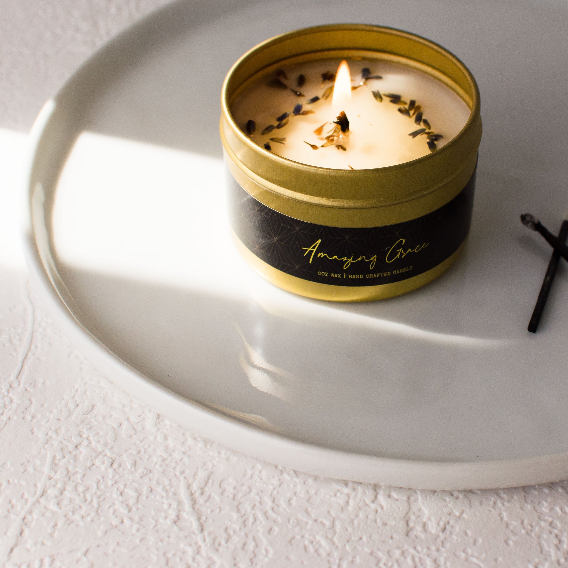 A top down shot of a gold 4oz tin soy candle with a black label named Amazing Grace with dried lavender buds on top of the melted wax in a clean white tray, made by Ebony & Ivory Candle Co in Lethbridge, Alberta, Canada