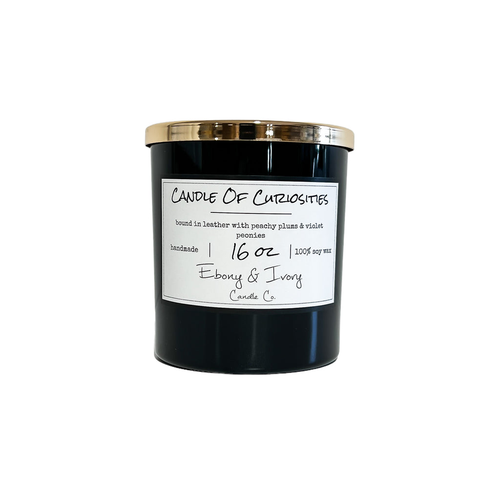 Black, sixteen ounces, Leather, peaches, plums, violets, and peonies scented soy wax candle with a gold lid and a white label that reads Candle of Curiosity