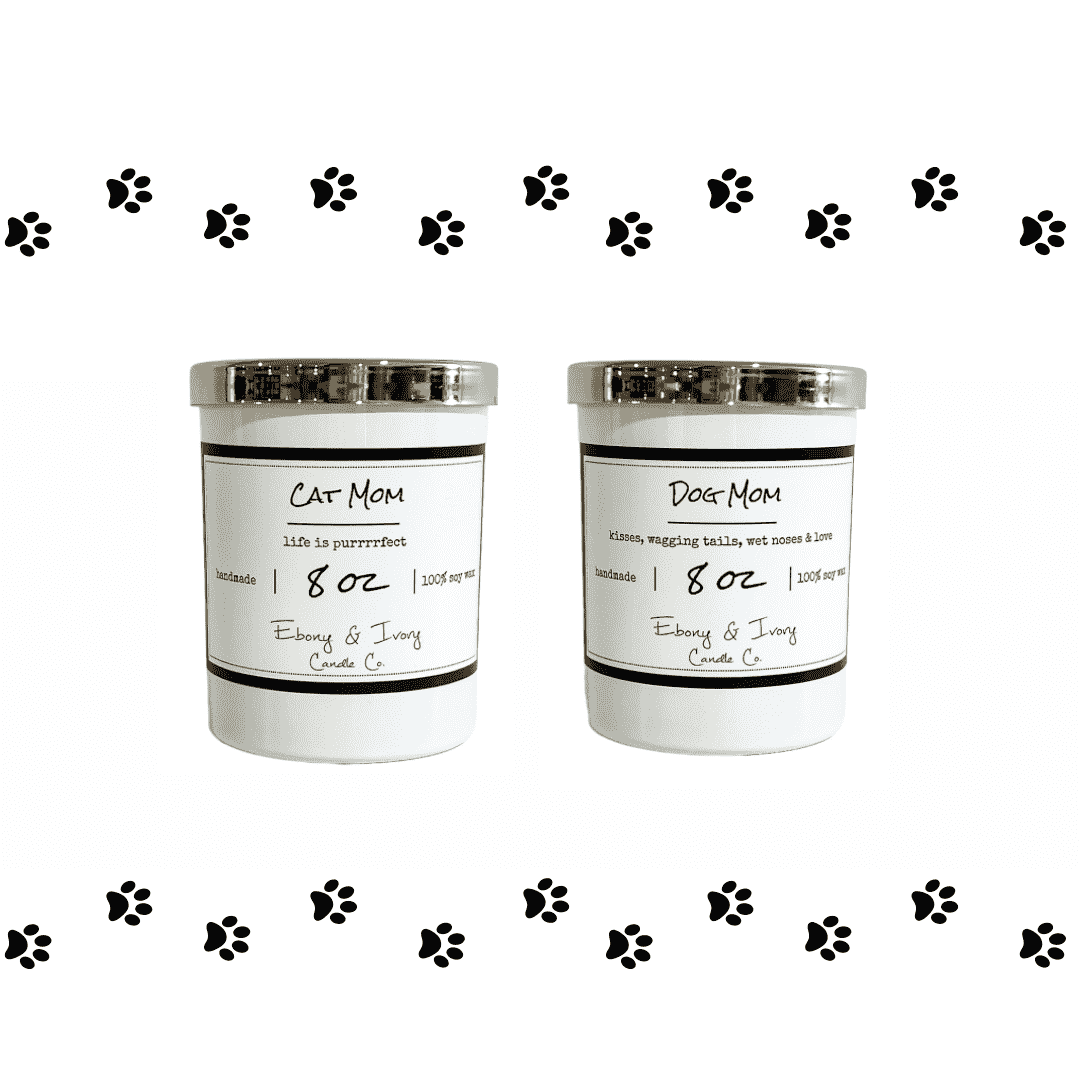 Two white 8oz soy candles with gold lids, one labeled 'Cat Mom' and the other 'Dog Mom', with black decorative paw prints around them. Made by ebony & ivory candle co in lethbridge alberta
