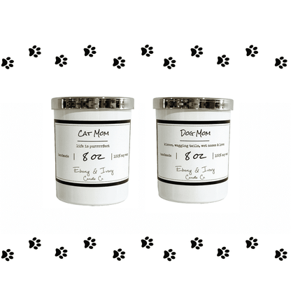 Two white 8oz soy candles with gold lids, one labeled 'Cat Mom' and the other 'Dog Mom', with black decorative paw prints around them. Made by ebony & ivory candle co in lethbridge alberta