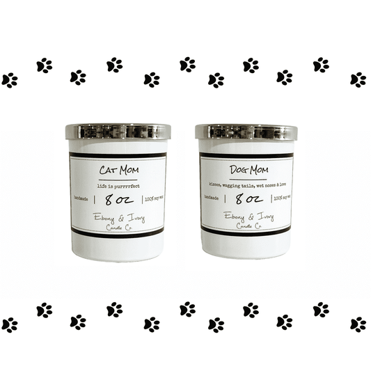 Two white 8oz soy candles with gold lids, one labeled 'Cat Mom' and the other 'Dog Mom', with black decorative paw prints around them. Made by ebony & ivory candle co in lethbridge alberta