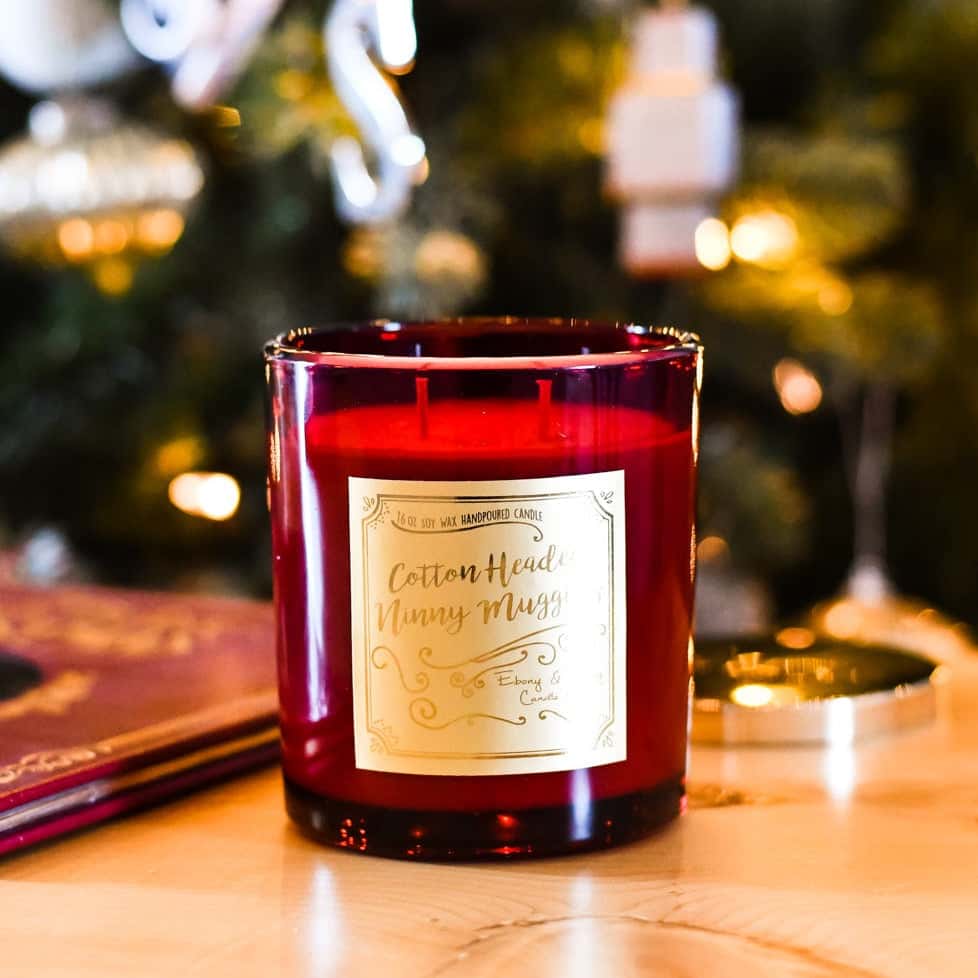 A red 16oz soy candle with a gold label named "cotton headed ninny muggins" sitting next to a christmas story book, with a christmas tree behind it.