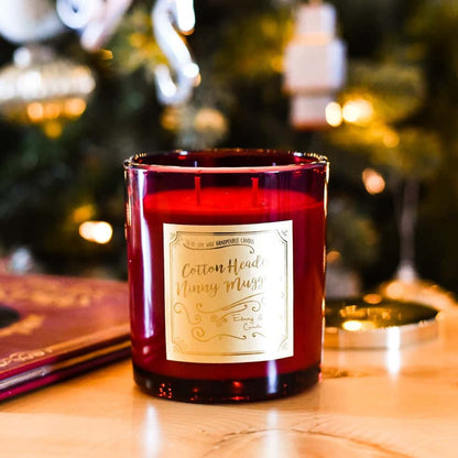 A red 16oz soy candle with a gold label named "cotton headed ninny muggins" sitting next to a christmas story book, with a christmas tree behind it.