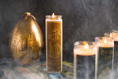 Tall 16oz clear glass jar soy candle with a green label named Butter Beer, on a stack of harry potter books with smoke billowing around, apart of the wizarding candle collection made by Ebony & Ivory Candle Co in Lethbridge, Alberta, Canada