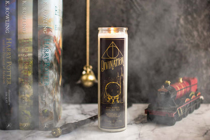 Tall 16oz clear glass jar soy candle with a green label named Divination, on a marble table with harry potter books, a wand, and the hogwarts train behind it, with smoke billowing around. apart of the wizarding candle collection made by Ebony & Ivory Candle Co in Lethbridge, Alberta, Canada