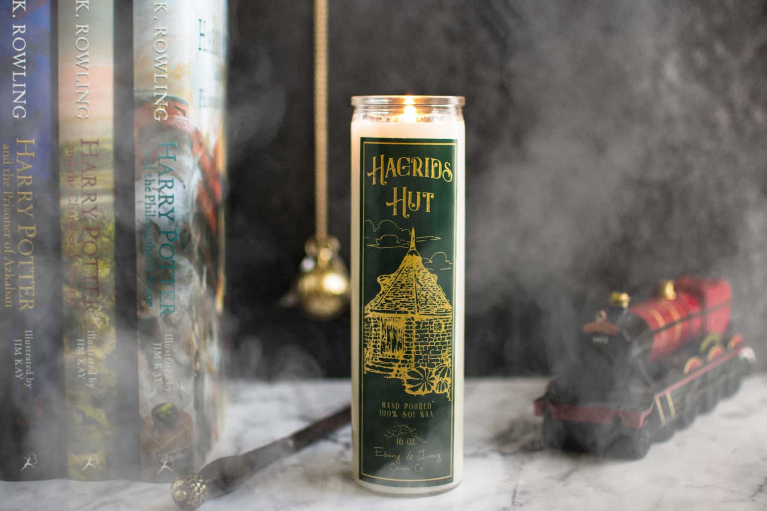 Tall 16oz clear glass jar soy candle with a green label named Hagrid's Hut, on a marble table with harry potter books, a wand, and the hogwarts train behind it, with smoke billowing around. apart of the wizarding candle collection made by Ebony & Ivory Candle Co in Lethbridge, Alberta, Canada