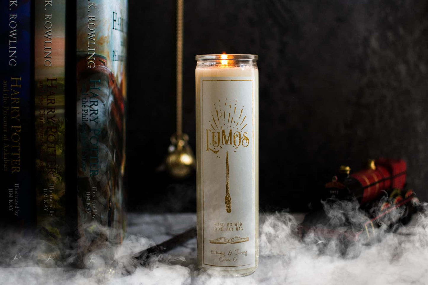 Tall 16oz clear glass jar soy candle with a green label named Lumos, on a marble table with harry potter books, a wand, and the hogwarts train behind it, with smoke billowing around. apart of the wizarding candle collection made by Ebony & Ivory Candle Co in Lethbridge, Alberta, Canada