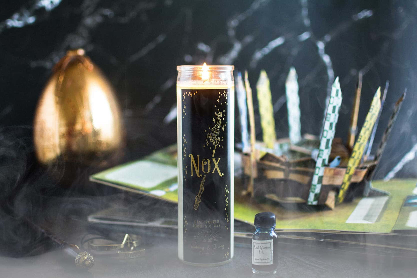 Tall 16oz clear glass jar soy candle with a green label named Nox, on a dark table with a harry potter pop up book and a wand behind it. apart of the wizarding candle collection made by Ebony & Ivory Candle Co in Lethbridge, Alberta, Canada