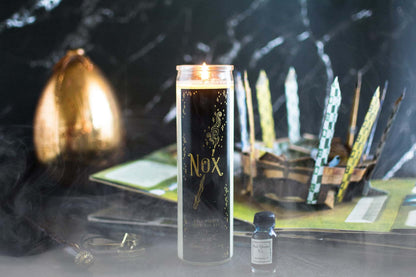 Tall 16oz clear glass jar soy candle with a green label named Nox, on a dark table with a harry potter pop up book and a wand behind it. apart of the wizarding candle collection made by Ebony & Ivory Candle Co in Lethbridge, Alberta, Canada
