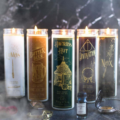 5 tall, clear, 16oz soy candles, one with a green label named Hagrid's Hut, one with a white label named Lumos, one with a gold label named Butter Beer, one with a black label named Nox, and one with a purple label named Divination. on a black table with Harry Potter themed items in front of them and smoke billowing around. All apart of the wizarding candle collection made by Ebony & Ivory Candle Co in Lethbridge, Alberta, Canada