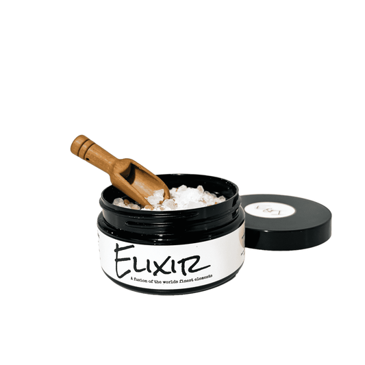 Black container with a white label that reads "elixir" and is filled with pink Himalayan salt, dead sea salt, epsom salt, and peppermint essential oils. A wooden spook is scoop some of the bath soak.