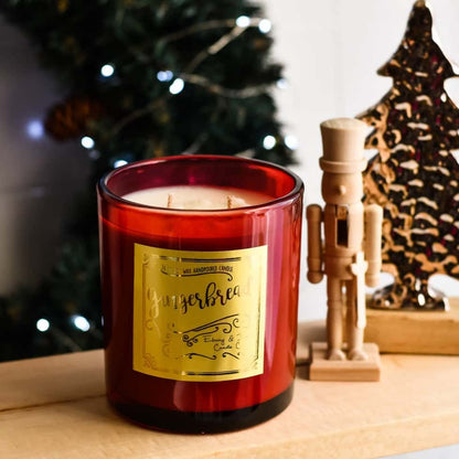 red 16oz soy christmas candle with a gold label named "gingerbread, sitting on a table with christmas lights and holiday decor.