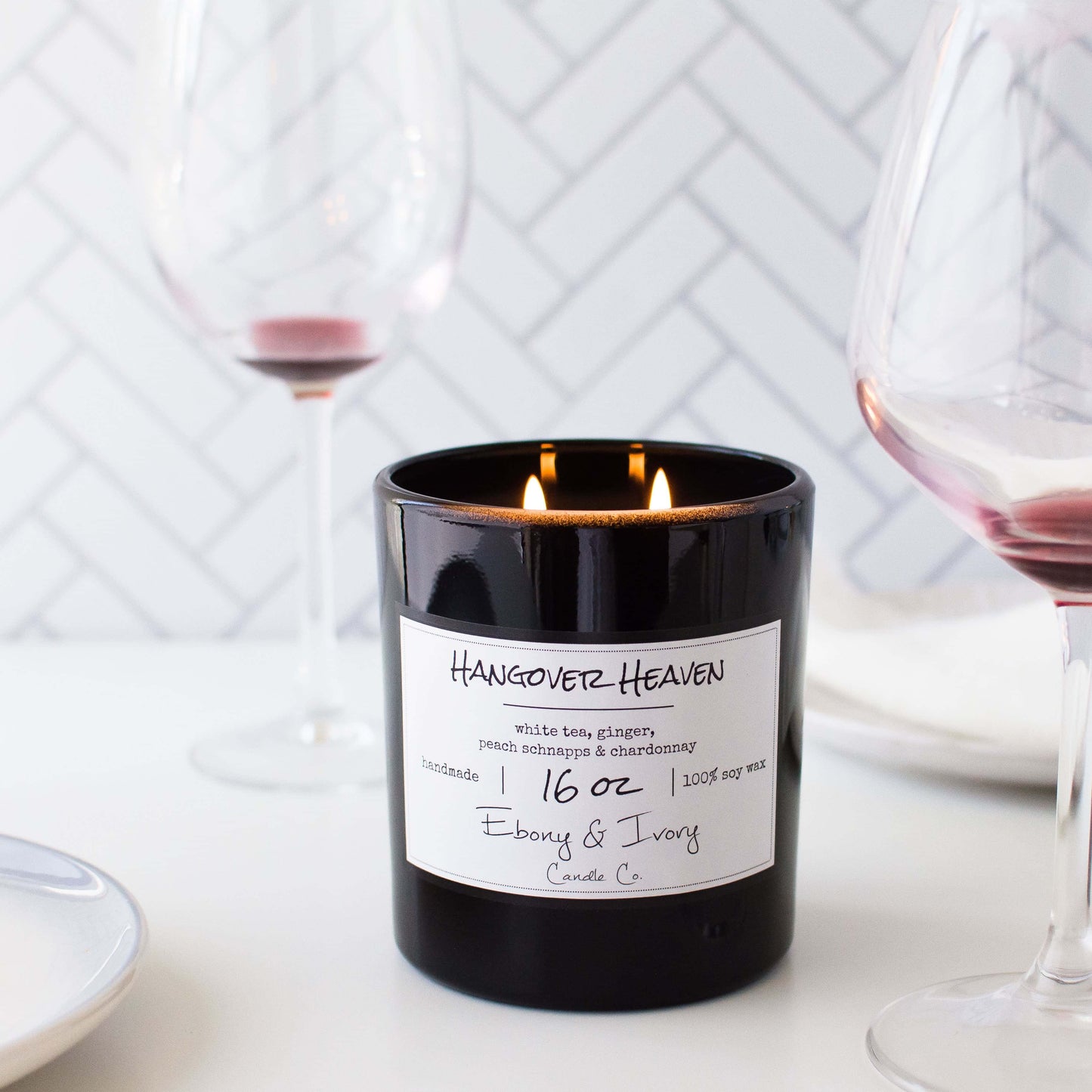 A black 16oz jar soy candle with a gold lid and a white label named Hangover Heaven, on a white table with wine glasses, made by Ebony & Ivory Candle Co in Lethbridge, Alberta, Canada