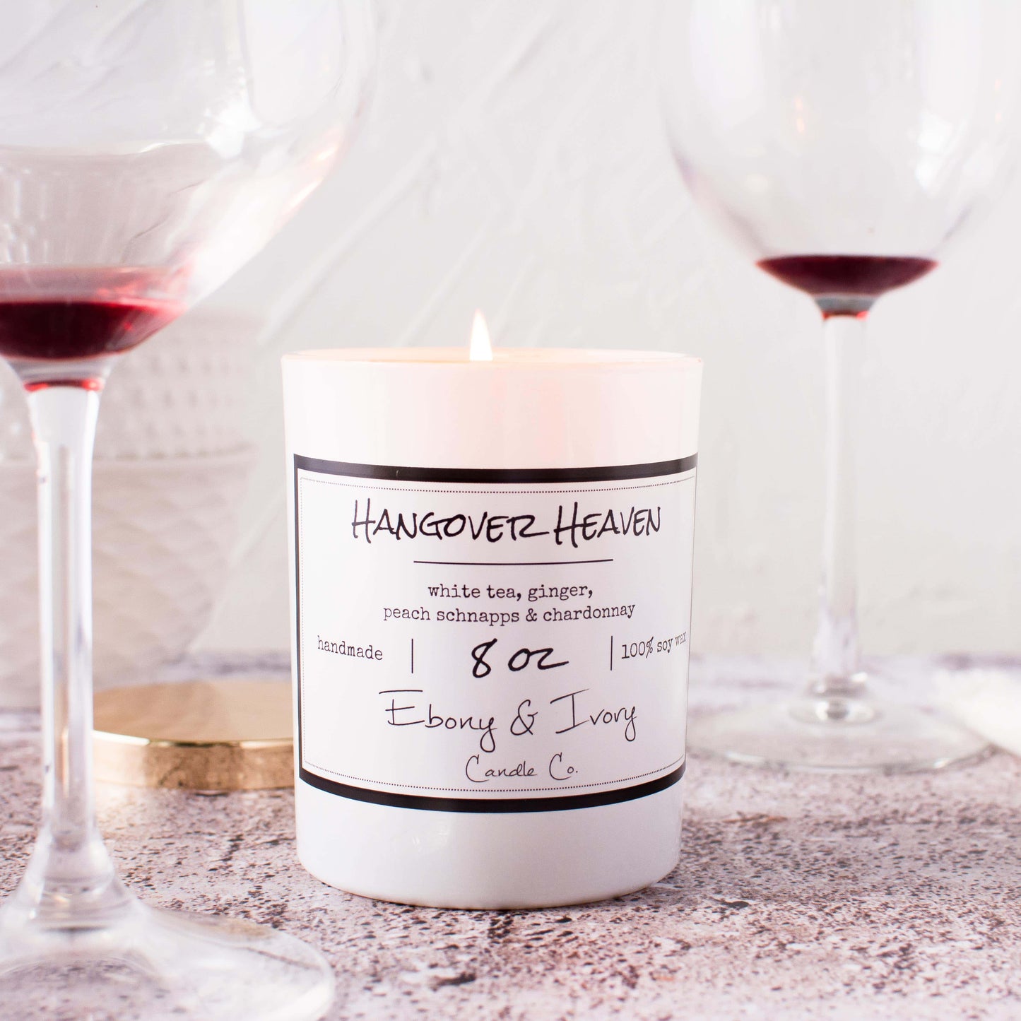 A white 8oz jar soy candle with a gold lid and a white label named Hangover Heaven, on a counter top with wine glasses, made by Ebony & Ivory Candle Co in Lethbridge, Alberta, Canada