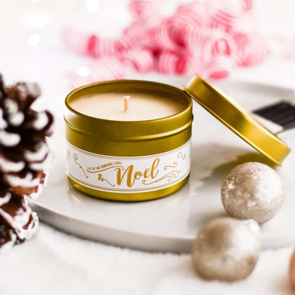 Gold 4oz, cedar, fir, cinnamon, clove, orange, and lemon scented soy wax candle with a white label and a gold lid named "noel" sitting on a white plate with christmas decorations