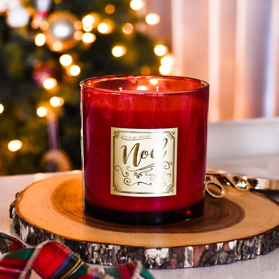 a red 64oz soy candle with a gold label named "noel" sitting on a wood surface with ribbon and a christmas tree with lights in the background