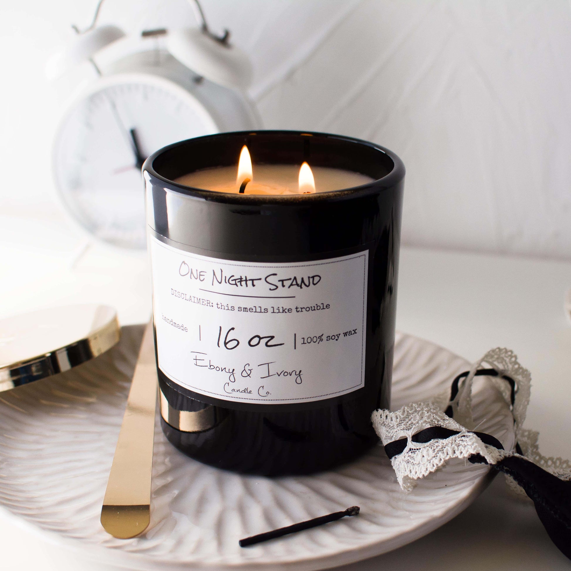 A black jar 16oz soy candle with a white label named One Night Stand, on a white dish with a black lacey tank top and a white alarm clock in the background, made by Ebony & Ivory Candle Co in Lethbridge, Alberta, Canada
