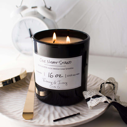 A black jar 16oz soy candle with a white label named One Night Stand, on a white dish with a black lacey tank top and a white alarm clock in the background, made by Ebony & Ivory Candle Co in Lethbridge, Alberta, Canada