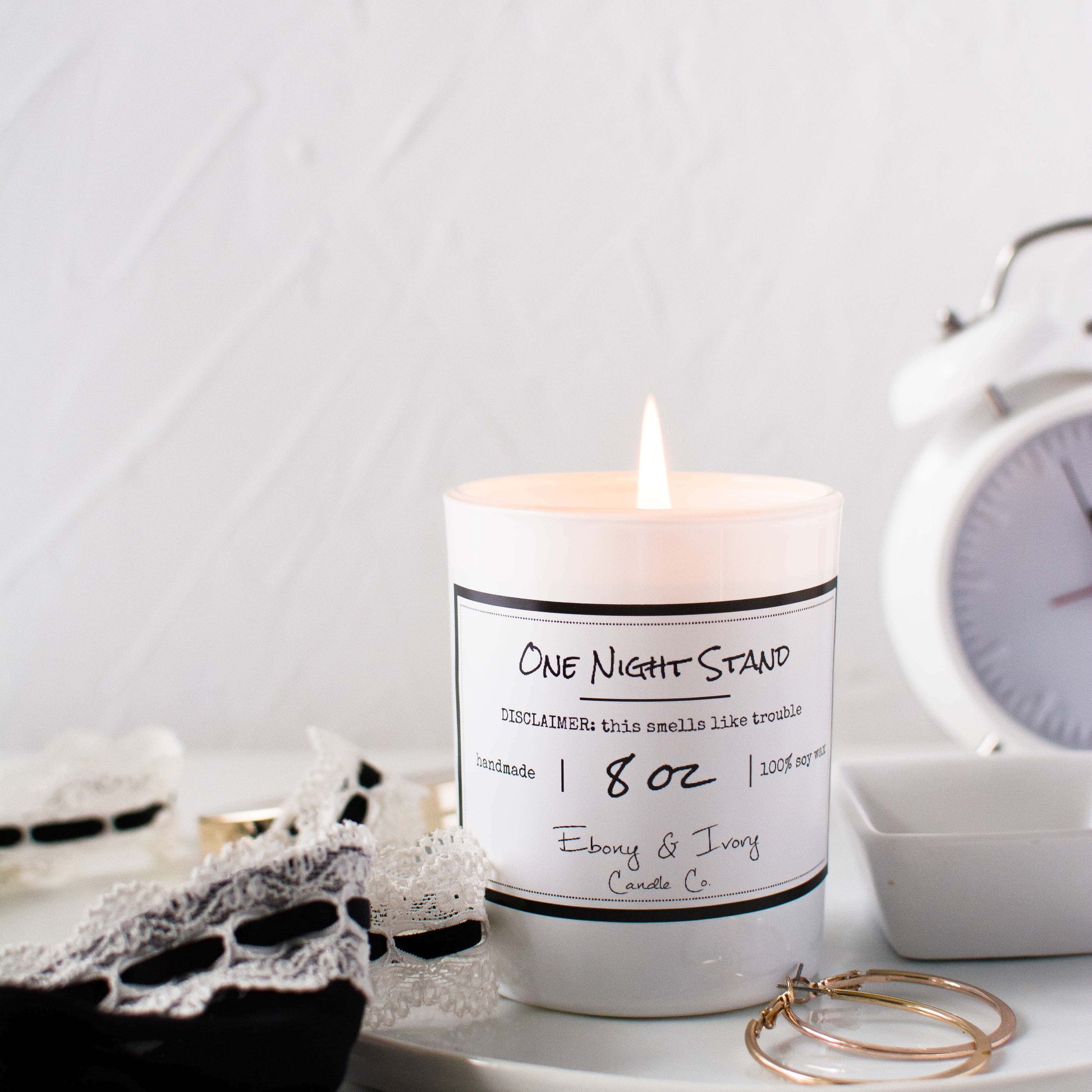 A white jar 8oz soy candle with a gold lid and a white label named One Night Stand, on a white night stand with gold hoop earrings, a black lacey tank top, and a white alarm clock in the background, made by Ebony & Ivory Candle Co in Lethbridge, Alberta, Canada