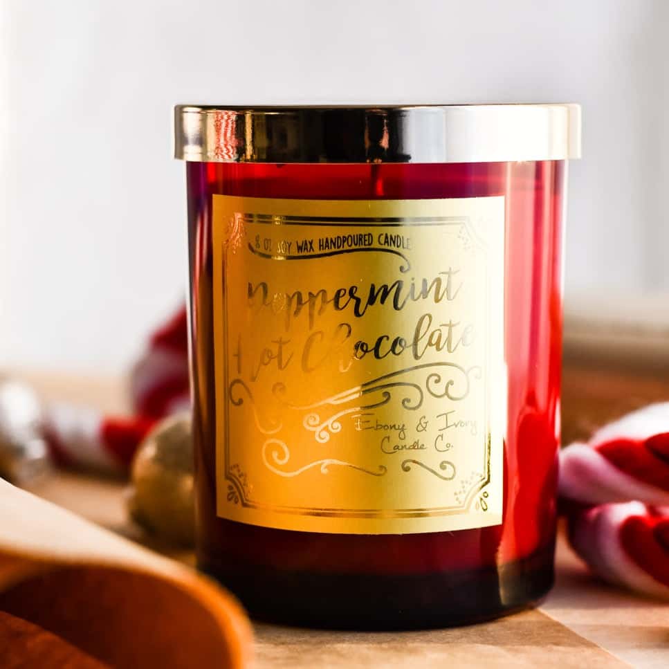 A red 8oz soy christmas candle with a gold lid and a gold label named "peppermint hot chocolate" sitting amongst holiday decorations