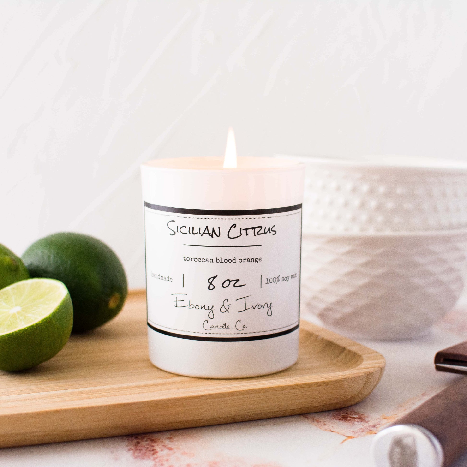 A white jar 8oz soy candle with a gold lid and a white label named Sicilian Citrus, on a light wood cutting board on top of a kitchen counter, with limes and white bowls behind it, made by Ebony & Ivory Candle Co in Lethbridge, Alberta, Canada