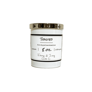 White, eight ounce, marshmallow, vanilla, and woodsy scented soy wax candle with a gold lid and a white label that reads Toasted